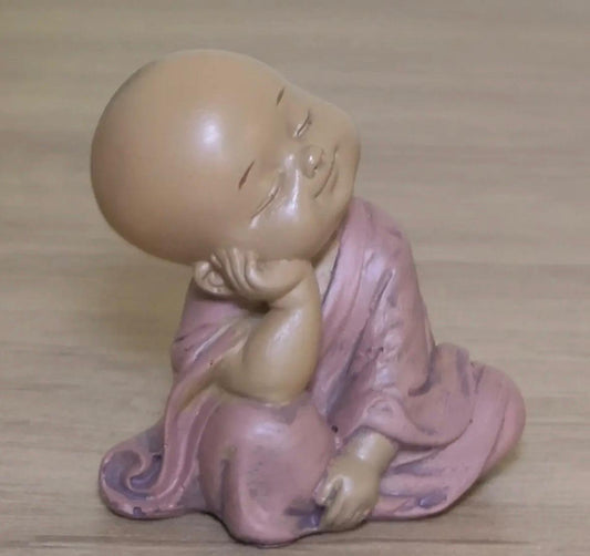 Figurine de petit Bouddha souriant en tenue rose, assis avec la tête appuyée sur une main. 