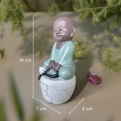 Figurine de petit moine assis en tenue vert clair, tenant un chapelet noir, avec dimensions indiquées : 14 × 7 × 6 cm.