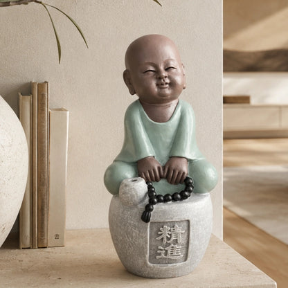 Figurine de petit moine souriant en tenue vert clair, assis sur un socle gris avec un chapelet noir, près de livres et d’un vase.