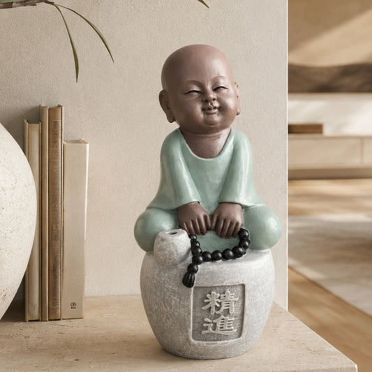 Figurine de petit moine souriant en tenue vert clair, assis sur un socle gris avec un chapelet noir, près de livres et d’un vase.
