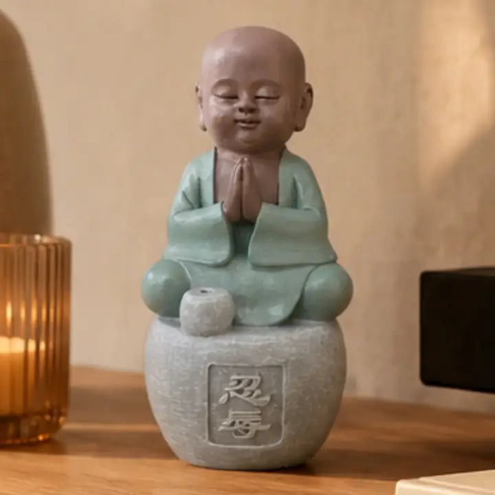 Figurine de petit moine assis en tenue vert clair, les mains jointes, sur un socle gris près d’une bougie.