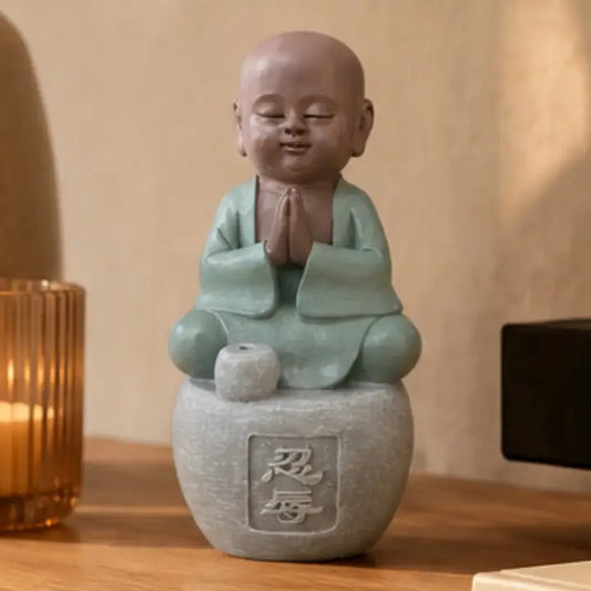 Figurine de petit moine assis en tenue vert clair, les mains jointes, sur un socle gris près d’une bougie.